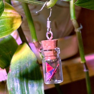 May include: A clear glass bottle earring with a cork stopper, containing a small origami crane in shades of red and purple. The earring hangs from a silver hook and is set against a backdrop of green leaves.