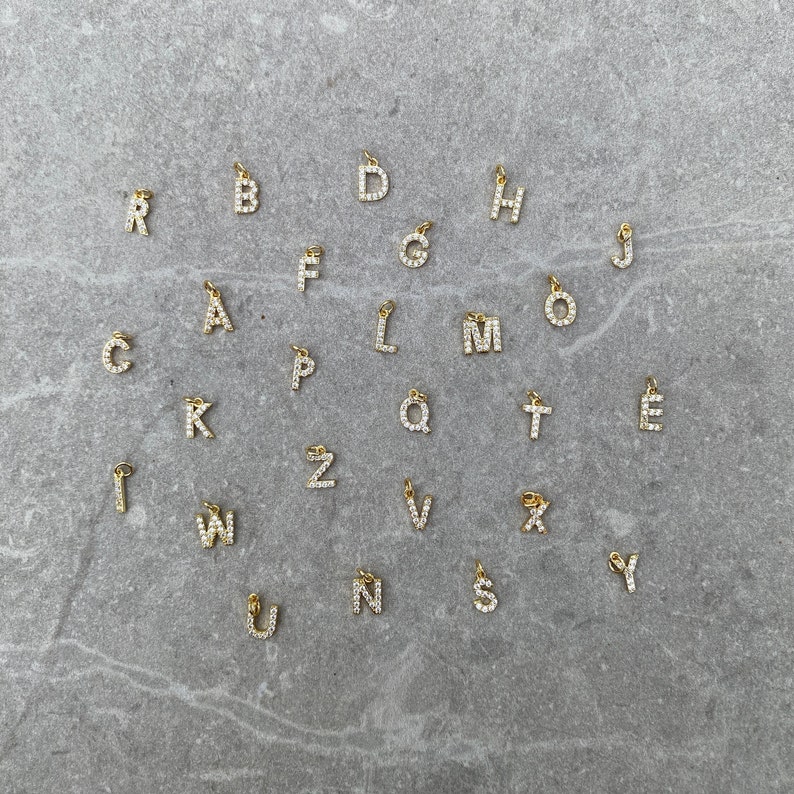 May include: A set of 26 gold letter charms, each letter is encrusted with small clear rhinestones. The charms are arranged in alphabetical order on a gray textured surface.