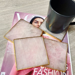 May include: Four square rose quartz coasters with gold edging. A black mug rests on one coaster. The coasters are on a magazine with the word "Vogue" visible. The magazine has the text "RACE & THE NAGA MUNCHE TALKS".