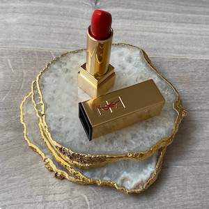 May include: A gold lipstick tube with a red lipstick bullet on top, sitting on a white agate coaster with gold trim.