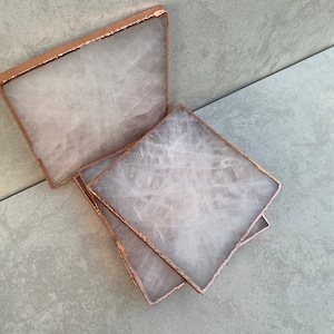 May include: Square coasters with a pale grey, translucent appearance and a copper-coloured trim. The coasters are made of a natural stone material and are arranged on a light grey surface.