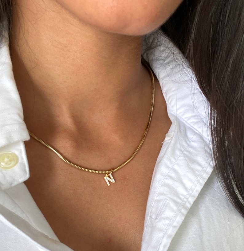 May include: A gold chain necklace with a small pendant in the shape of the letter N, adorned with sparkling clear stones.