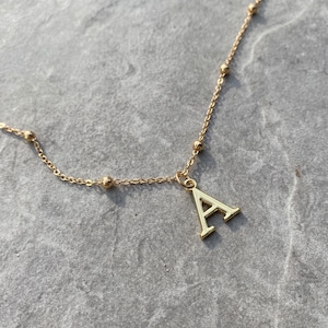 Puede incluir: Un collar de cadena dorada con un pequeño dije de bola dorada y un dije de letra A dorada más grande.