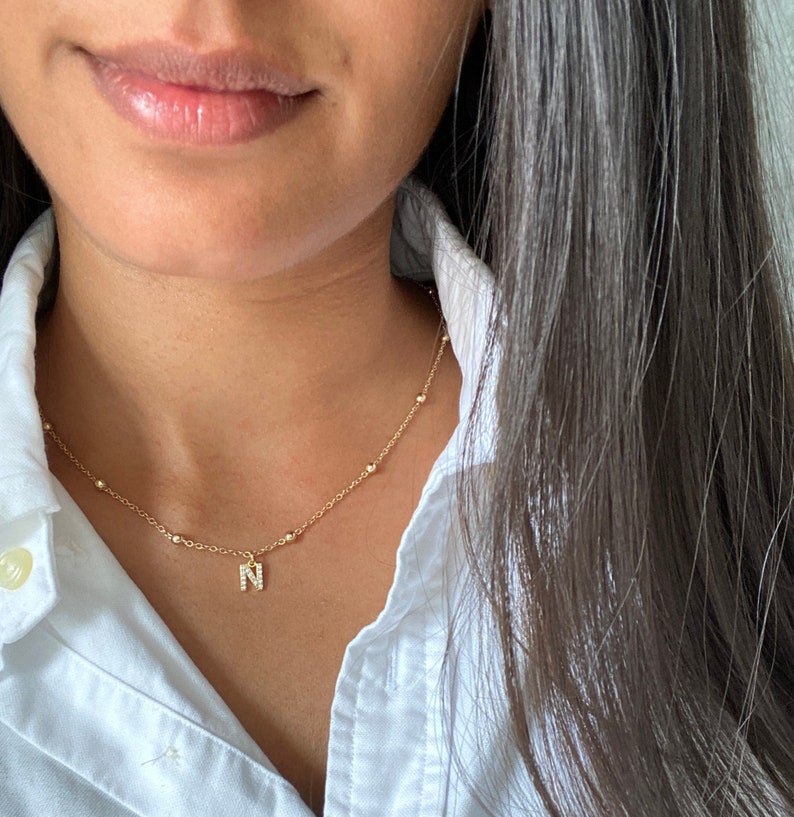 May include: A gold chain necklace with a small pendant featuring the letter 'N' encrusted with tiny clear stones.