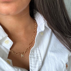 May include: A gold chain necklace with a small letter 'N' charm. The charm is set with small clear stones. The necklace is worn by a person wearing a white collared shirt.