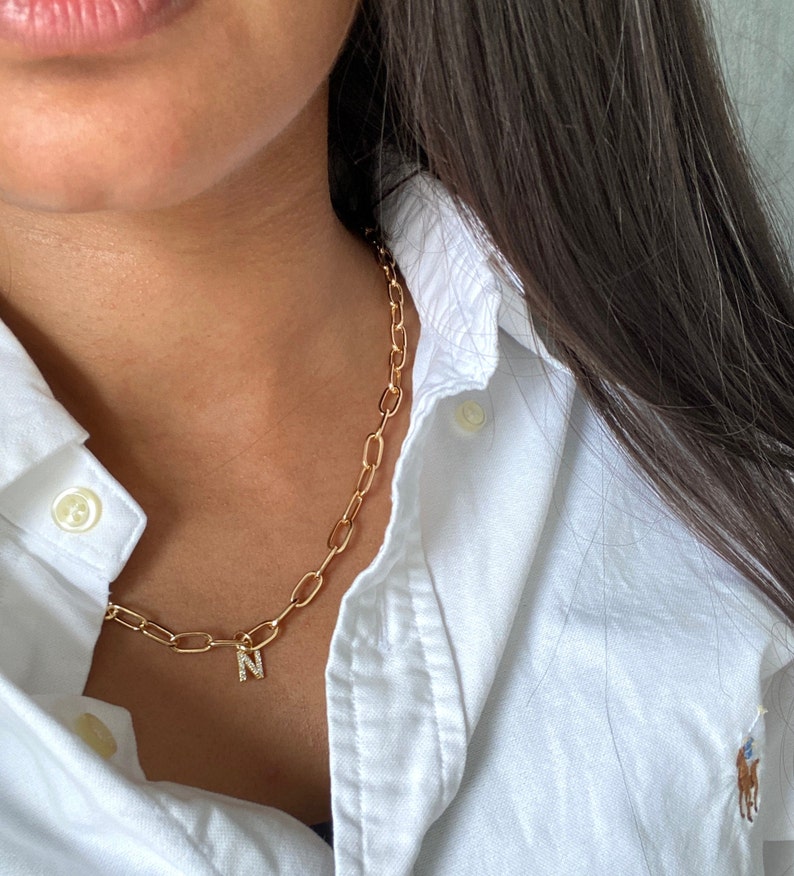 May include: A gold chain necklace with a small letter 'N' charm. The charm is set with small clear stones. The necklace is worn by a person wearing a white collared shirt.