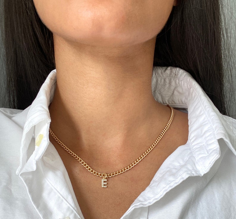 May include: A gold chain necklace with a small pendant in the shape of the letter "E" with small clear stones. The necklace is worn by a person with long dark hair.