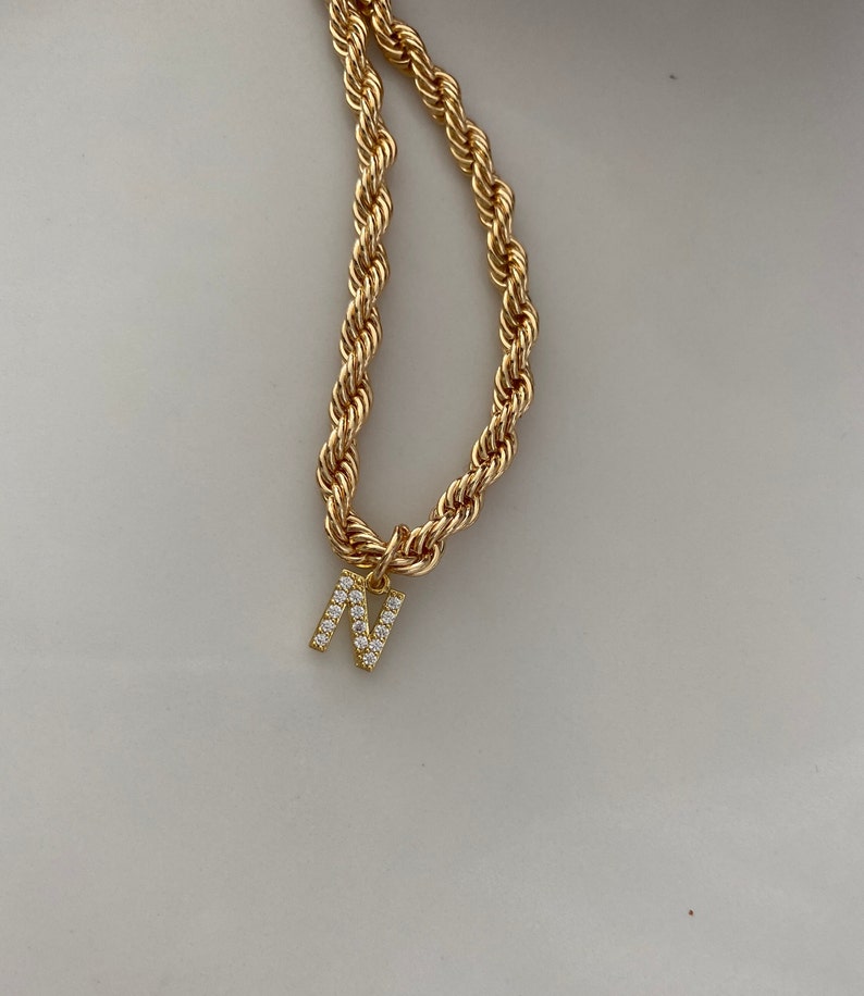 May include: A gold rope chain necklace with a gold letter N pendant encrusted with clear rhinestones.
