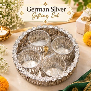 May include: A circular silver-toned metal tray with an ornate, scalloped edge featuring four matching small, round bowls inside. The tray and bowls have intricate cut-out patterns. A label above reads German Sliver Gifting Set.
