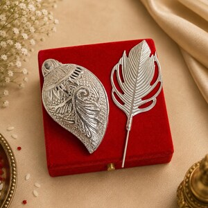 Puede incluir: Dos objetos decorativos plateados sobre una caja de terciopelo roja. Uno es una forma de concha ornamentada y detallada, y el otro es un diseño de pluma. El fondo es beige neutro con acentos florales, lo que sugiere un artículo decorativo o de regalo.