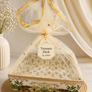 May include: A rectangular gift tray featuring a white base with a colorful elephant and architectural print, wrapped in sheer gold-trimmed tulle netting. A cream-colored tag attached to the top reads Treasure Deck by sakshi in black text.