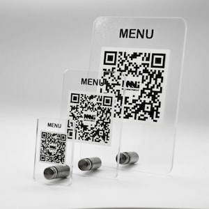May include: Three clear acrylic menu holders of varying sizes. Each holder features a QR code and the word "MENU" in black. The holders are supported by small, cylindrical metal bases, creating a modern and minimalist design.