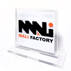 K&ouml;nnte beinhalten: Transparenter Acrylschildhalter mit dem Text "MALI FACTORY" in Schwarz und Orange. Der Halter hat einen rechteckigen Sockel und einen Schlitz f&uuml;r eine gedruckte Karte. Das Logo zeigt ein stilisiertes "NNN"-Design.