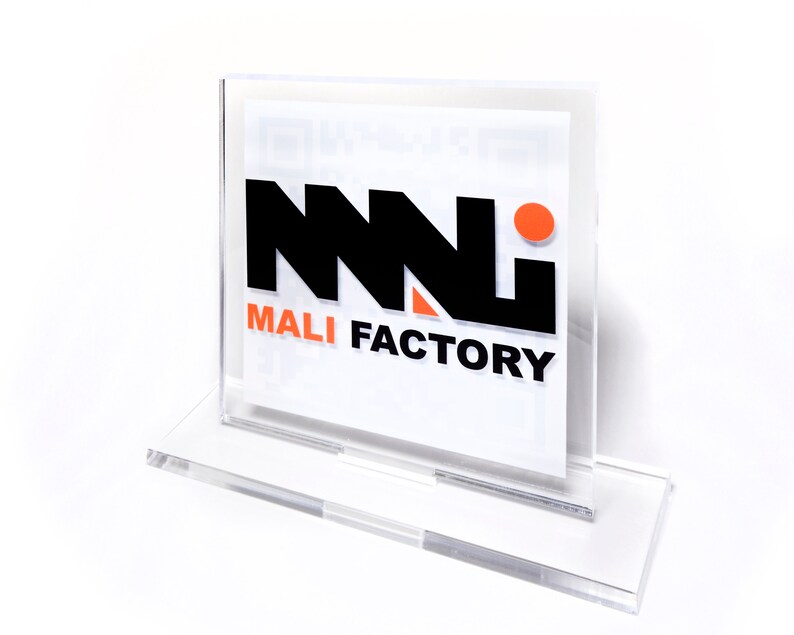 K&ouml;nnte beinhalten: Transparenter Acrylschildhalter mit dem Text "MALI FACTORY" in Schwarz und Orange. Der Halter hat einen rechteckigen Sockel und einen Schlitz f&uuml;r eine gedruckte Karte. Das Logo zeigt ein stilisiertes "NNN"-Design.