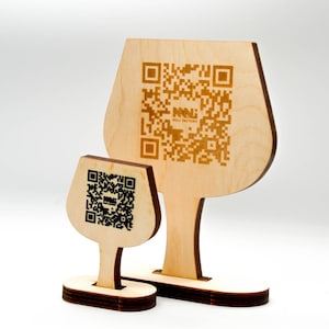 Puede incluir: Dos letreros de madera con forma de copa de vino con códigos QR. El letrero más grande tiene un tallo y una base, y el letrero más pequeño tiene un diseño similar. Los letreros son de madera clara y presentan el texto "MALI FACTORY".