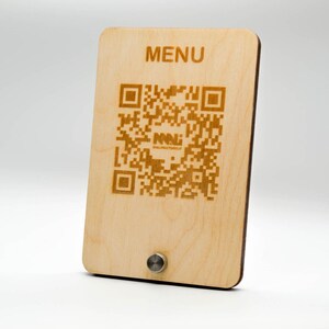 May include: A rectangular wooden menu board with rounded corners. The word "MENU" is at the top. A QR code and the text "NNL MALUFACTORY.IT" are laser-etched on the light-colored wood. A small, silver-colored metal knob is at the bottom.