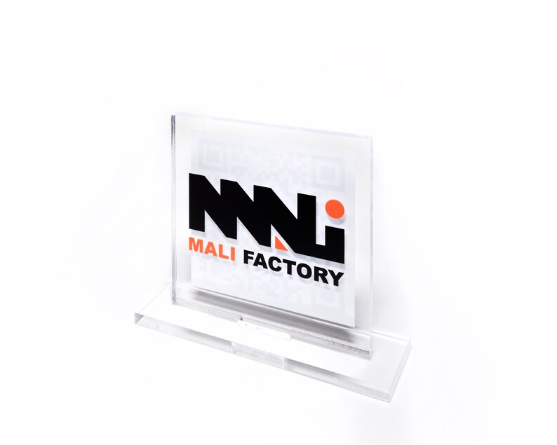 K&ouml;nnte beinhalten: Ein klarer Acryl-Displayst&auml;nder mit dem Text "MALI FACTORY" in Schwarz und Orange. Der St&auml;nder ist f&uuml;r eine Karte oder ein Schild konzipiert und hat eine quadratische Form mit einem Sockel f&uuml;r Stabilit&auml;t. Der Hintergrund ist wei&szlig;.
