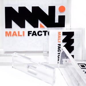 K&ouml;nnte beinhalten: Klare Acryl-Displayst&auml;nder mit bedruckten Eins&auml;tzen. Die Eins&auml;tze zeigen den Text "MALI FACTORY" in Schwarz und Orange sowie ein stilisiertes Logo. Einige Eins&auml;tze enthalten QR-Codes. Die St&auml;nder sind zur Aufnahme der Drucksachen konzipiert.