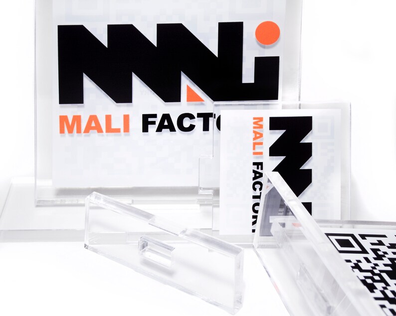 K&ouml;nnte beinhalten: Klare Acryl-Displayst&auml;nder mit bedruckten Eins&auml;tzen. Die Eins&auml;tze zeigen den Text "MALI FACTORY" in Schwarz und Orange sowie ein stilisiertes Logo. Einige Eins&auml;tze enthalten QR-Codes. Die St&auml;nder sind zur Aufnahme der Drucksachen konzipiert.