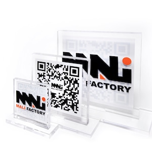 Puede incluir: Expositores de acrílico transparente de varios tamaños, cada uno con un código QR y el logotipo "MALI FACTORY" en negro y naranja. Los expositores están dispuestos en una configuración escalonada sobre un fondo blanco.