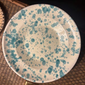 Op de afbeelding: Een rond keramieken bord met een glanzende witte basis voorzien van een gespikkeld patroon van diverse maten blauwgroene verfspatten over het oppervlak. Het bord heeft een brede, platte rand en een gladde, reflecterende afwerking.