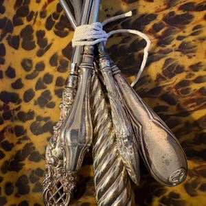 Puede incluir: Un conjunto de utensilios de plata antiguos atados con hilo blanco. Los cubiertos tienen mangos ornamentados con varios diseños, incluyendo patrones florales y trenzados. El fondo presenta un patrón de carey.