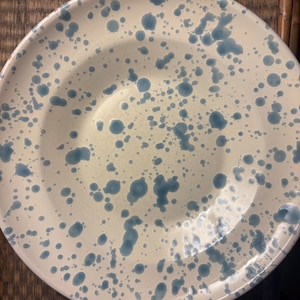 Puede incluir: Un plato redondo de cerámica con una base de color crema que presenta un patrón de manchas azules. El diseño consiste en salpicaduras de pintura azul de varios tamaños distribuidas por toda la superficie del plato, incluyendo el borde ancho.