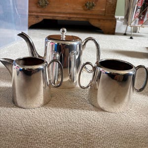 Puede incluir: Un juego de té plateado que incluye una tetera, una lechera y un azucarero. La tetera tiene un pico curvo y una tapa con un pequeño pomo. La lechera y el azucarero tienen asas curvas. El juego se exhibe sobre una alfombra de color neutro.