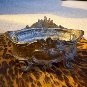 May include: An antique bronze-toned decorative bowl with ornate scrollwork and a central cartouche. The bowl has a shallow basin and is supported by small feet. The overall design is reminiscent of vintage decor.