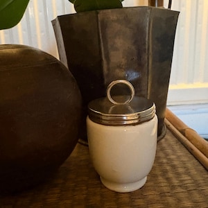 May include: A small, off-white ceramic jar with a smooth, rounded body and a flared base. The jar features a polished silver-tone metal lid that is topped with a circular ring handle, providing a simple and functional design for storage.