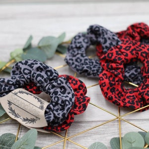 Könnte beinhalten: Zwei Sätze Scrunchies im Leopardenmuster. Ein Satz ist grau und schwarz, der andere ist rot und schwarz. Die Scrunchies befinden sich auf einem goldenen Metallständer mit einem Holzschild, auf dem "Scrunchies" steht.