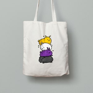 May include: A white tote bag with a design of four stacked cartoon bunnies. The bunnies are colored yellow, white, purple, and black, representing the colors of the non-binary pride flag.
