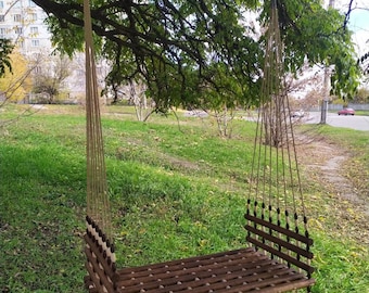Op de afbeelding: Een houten schommel die aan een tak van een boom hangt. De schommel is gemaakt van houten latten en heeft een touwophangingssysteem. De schommel hangt boven een grasveld.