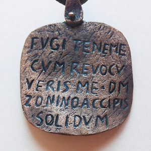 Puede incluir: Un colgante de bronce con un cordón negro. El colgante es rectangular y tiene una inscripción en latín: "FVGI TENEME CVM REVOCV VERIS ME DM ZONINO ACCIPIS SOLIDVM".