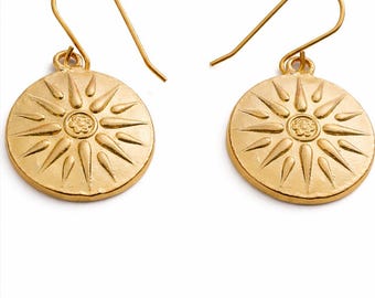 May include: A pair of gold-colored earrings. Each earring features a circular pendant with a sunburst design. The sunburst has pointed rays extending from a central floral-like design. The earrings have a simple hook closure.