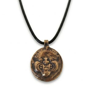 May include: A bronze-coloured pendant necklace. The circular pendant features a detailed face design and hangs from a black cord. The background is plain white.