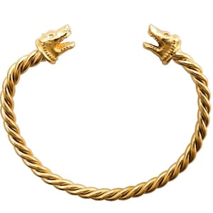 Op de afbeelding: Een goudkleurige, open manchetarmband met een gedraaid touwontwerp. De uiteinden van de armband zijn gebeeldhouwd in de hoofden van grommende wolven. De armband is een opvallend sieraad.