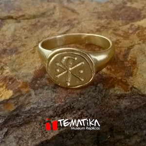 Puede incluir: Un anillo dorado con una cara ovalada que presenta un emblema detallado. El anillo est&aacute; sobre una superficie marr&oacute;n texturizada. El texto "TEMATIKA Museum Replicas" es visible en la parte inferior de la imagen.