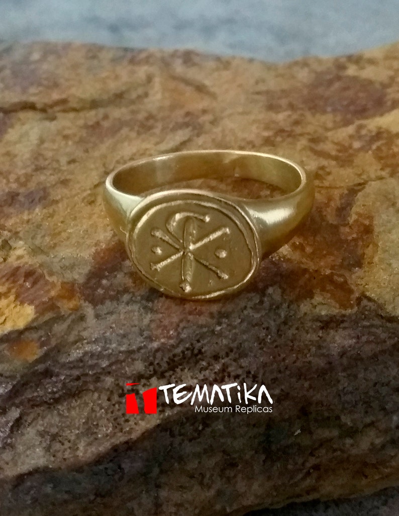 Puede incluir: Un anillo dorado con una cara ovalada que presenta un emblema detallado. El anillo est&aacute; sobre una superficie marr&oacute;n texturizada. El texto "TEMATIKA Museum Replicas" es visible en la parte inferior de la imagen.