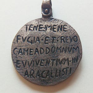 Könnte beinhalten: Ein runder, bronzener Anhänger mit einer Schlaufe oben. Der Anhänger trägt eine Inschrift, die lautet: "TENEMENE FVGIATREVOCAMEADDOMNVMEVIVVENTIMINARACALISTI".