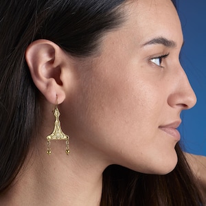 Puede incluir: Pendientes de aro dorados con un dise&ntilde;o geom&eacute;trico y peque&ntilde;as cuentas redondas. Los pendientes son usados por una persona con cabello largo y oscuro.