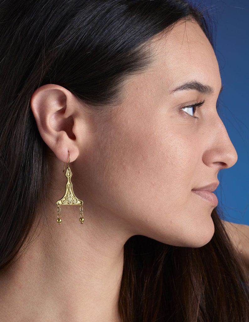 Puede incluir: Pendientes de aro dorados con un dise&ntilde;o geom&eacute;trico y peque&ntilde;as cuentas redondas. Los pendientes son usados por una persona con cabello largo y oscuro.