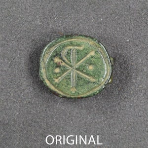 Puede incluir: Un artefacto redondo de bronce antiguo con una p&aacute;tina verde. El dise&ntilde;o presenta un s&iacute;mbolo Chi-Rho dentro de un c&iacute;rculo, con dos peque&ntilde;os puntos. La palabra "ORIGINAL" est&aacute; impresa en blanco debajo.