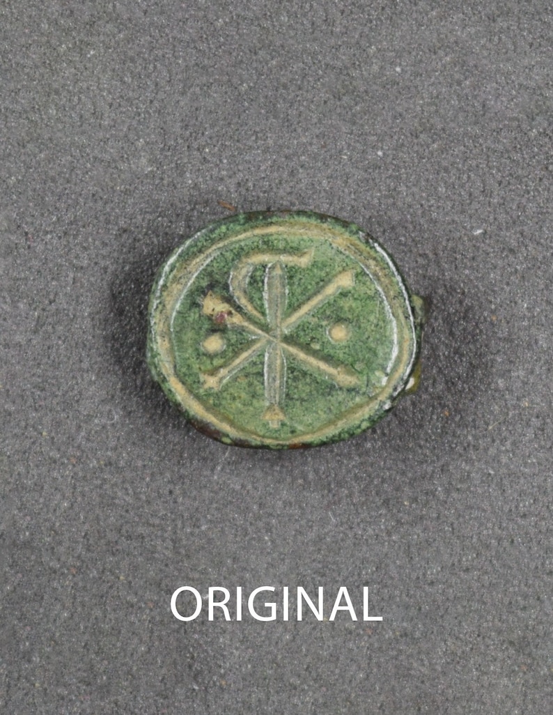 Puede incluir: Un artefacto redondo de bronce antiguo con una p&aacute;tina verde. El dise&ntilde;o presenta un s&iacute;mbolo Chi-Rho dentro de un c&iacute;rculo, con dos peque&ntilde;os puntos. La palabra "ORIGINAL" est&aacute; impresa en blanco debajo.