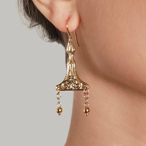 Puede incluir: Pendientes de aro dorados con un dise&ntilde;o geom&eacute;trico y una peque&ntilde;a perla al final de cada cadena.