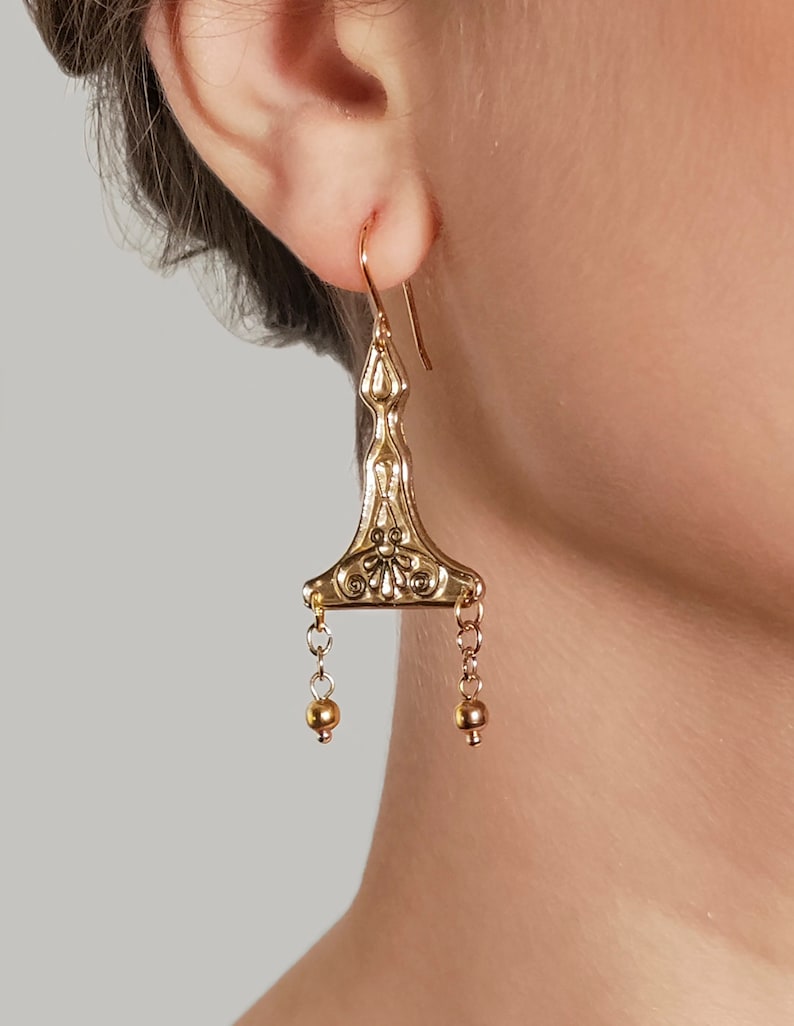 Puede incluir: Pendientes de aro dorados con un dise&ntilde;o geom&eacute;trico y una peque&ntilde;a perla al final de cada cadena.