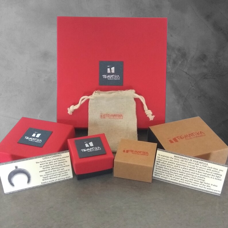 Puede incluir: Una caja roja con una etiqueta negra que dice "TEMATIKA" y una caja roja m&aacute;s peque&ntilde;a con una etiqueta negra que dice "TEMATIKA" encima de una caja marr&oacute;n con una etiqueta negra que dice "TEMATIKA". Hay una peque&ntilde;a bolsa blanca con una etiqueta negra que dice "TEMATIKA" encima de la caja roja. Las cajas est&aacute;n sobre un fondo gris. El texto en la tarjeta dice: "LUNULA EARRING The lunula was a hanging amulet worn by Greek women. Its properties are fertility and healing in addition to offering protection against the evil eye. The amulet was inspired by the original in the British Museum of London. Replica made of sterling silver."