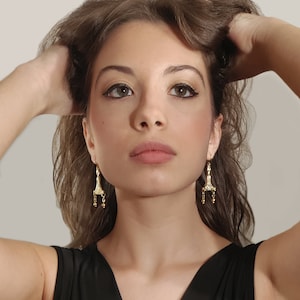 Puede incluir: Una mujer con el pelo largo y casta&ntilde;o lleva pendientes de oro con un dise&ntilde;o detallado. Los pendientes tienen forma de l&aacute;grima con una peque&ntilde;a cadena y cuentas que cuelgan de la parte inferior.