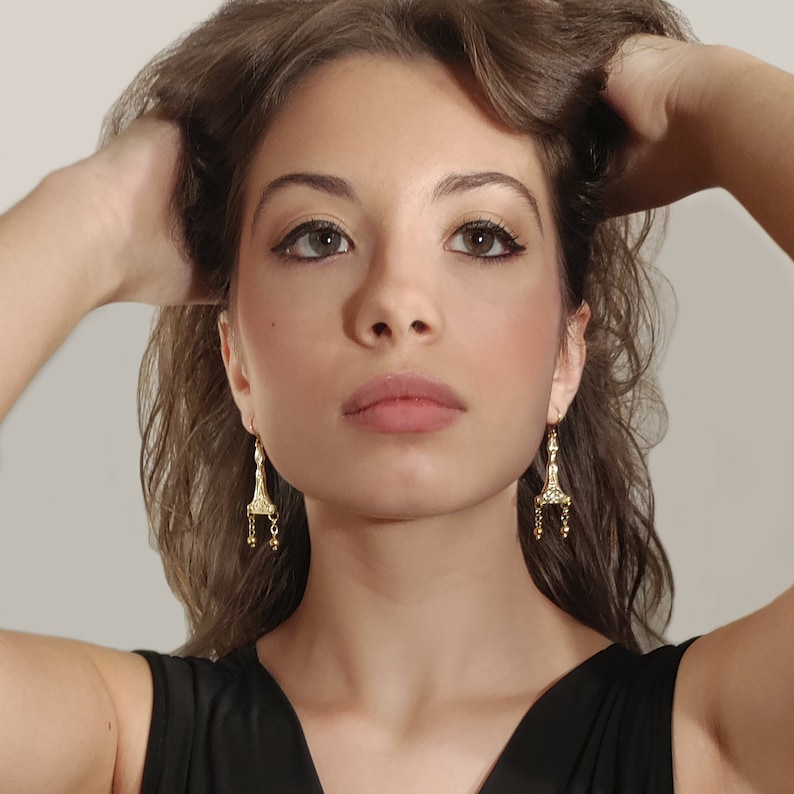 Puede incluir: Una mujer con el pelo largo y casta&ntilde;o lleva pendientes de oro con un dise&ntilde;o detallado. Los pendientes tienen forma de l&aacute;grima con una peque&ntilde;a cadena y cuentas que cuelgan de la parte inferior.