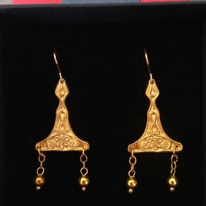 Puede incluir: Un par de pendientes de oro con un dise&ntilde;o triangular y cuentas colgantes. Los pendientes est&aacute;n en una caja negra con el texto "Tematika Museum Replicas".